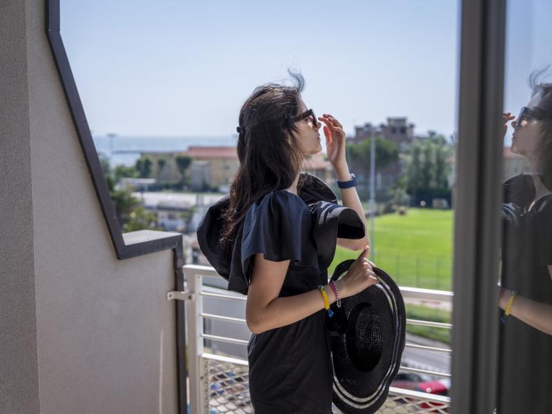 Donna con occhiali da sole e cappello, riflessa in una finestra su un balcone.